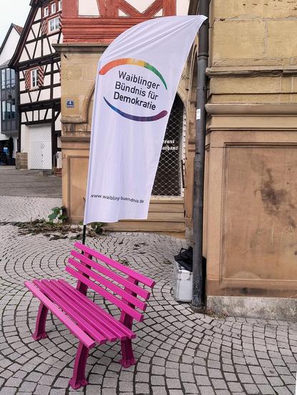 Die Pinke Bank dahinter eine Beach Flag mit dem runden regenbogenfarbenen Logo des Waiblinger Bündnis für Demokratie vor Altstadthäusern und Kopfsteinpflaster.