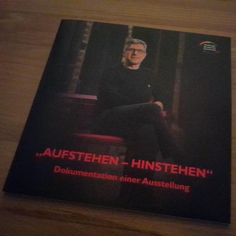 Titelseite der Dokumentation "AUFSTEHEN - HINSTEHEN" ein Mann sitzt auf der Pinken Bank der Demokratie.